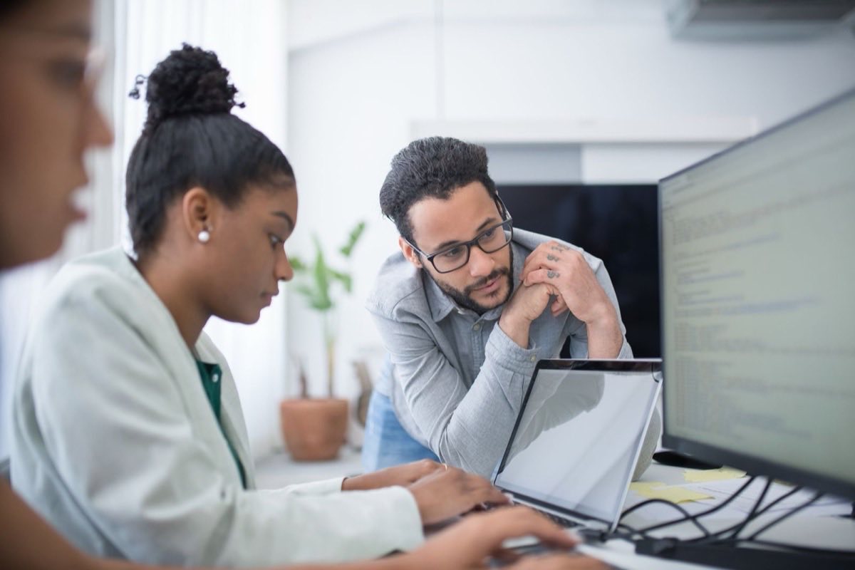 Two engineers leaning over a laptop and monitor together, reviewing code in a bright office