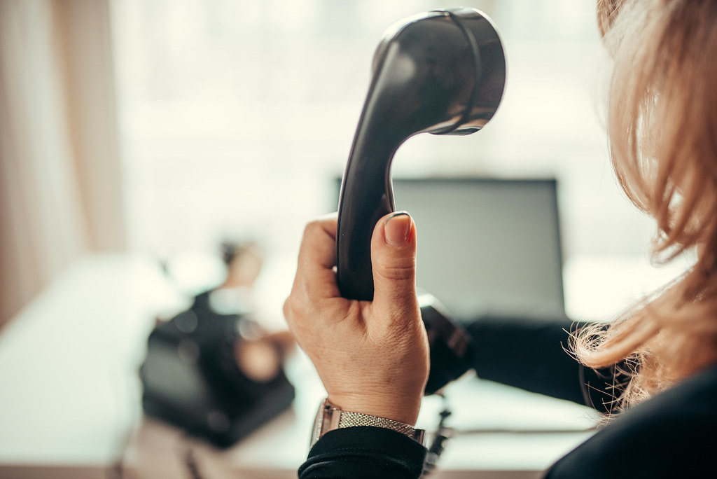 Close-up over-the-shoulder shot of a hand holding a black telephone receiver to make a call, laptop softly blurred in the warm light behind
