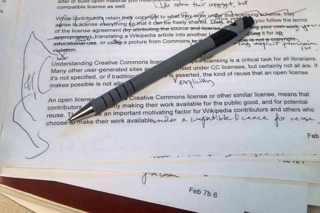 A printed manuscript page covered in handwritten corrections and marginal notes, a ballpoint pen lying across it on a stack of marked-up papers