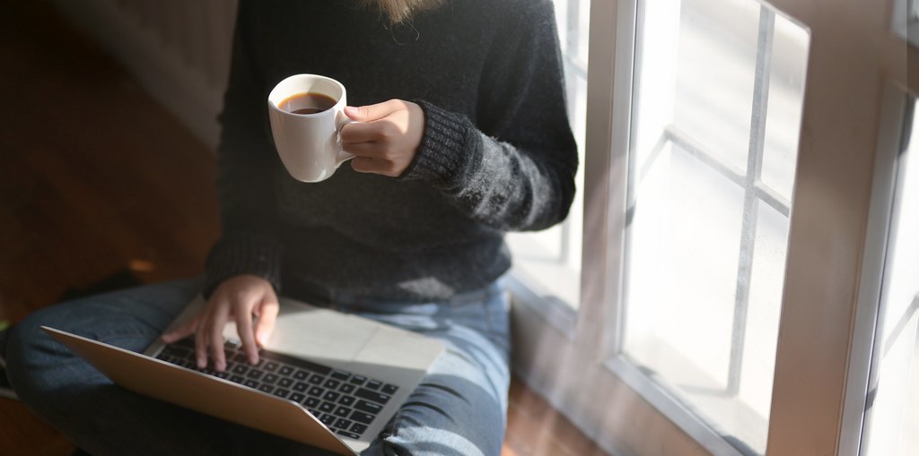 Woman by a sunlit window, coffee cup in hand, typing on a laptop resting on her lap